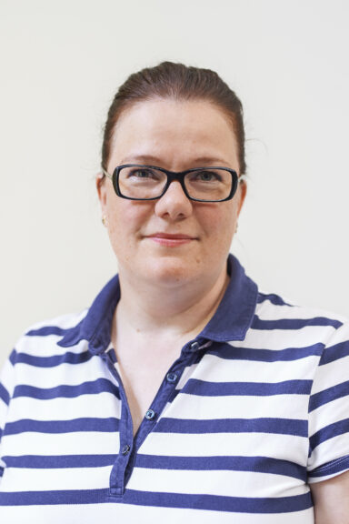 A woman looking friendly into the camera. She wears glasses. Her brown haire ist put in a ponytail. She is wearing a blue and white polo shirt.