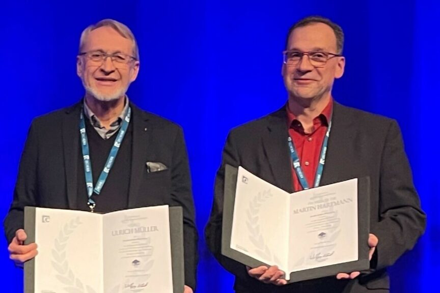 Two male senior scientists smiling in the camera, showing theier Award Certificate.