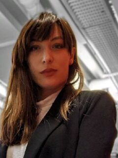 A woman in her 30th, with long brown/blond hair, dressed in a black suit, looking down in the camera selfconfident.